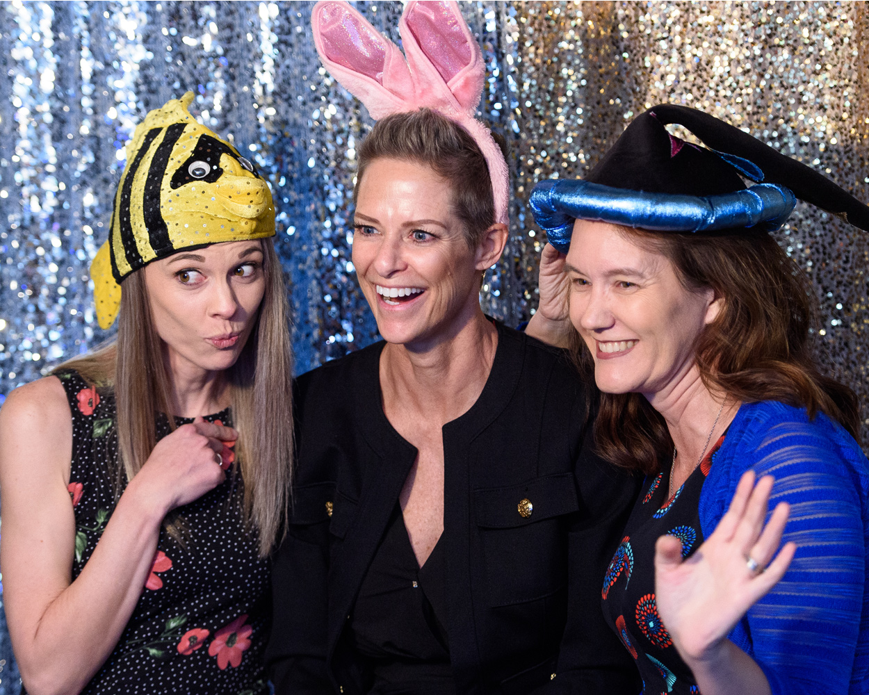 Three women faculty members smile in front of a glittery backdrop while wearing props including bunny ears